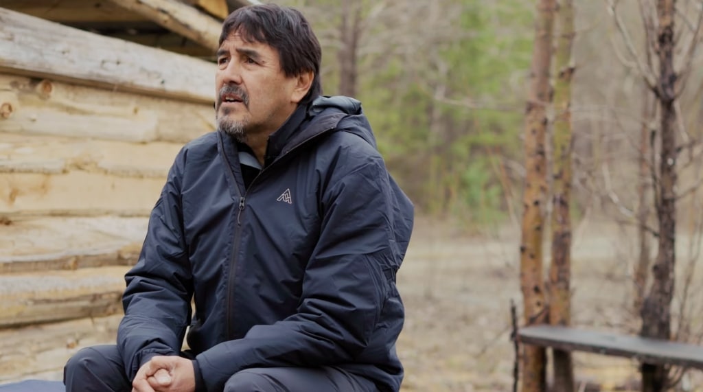 A man is sitting in the center of the frame. He is wearing dark, outdoor clothing and has his hands clasped. In the background is a wood cabin wall and some trees. 
